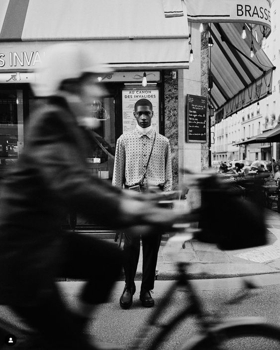 Stylishly dressed man standing still on a city street as a blurred cyclist passes by, black and white urban photo.