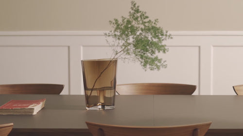 Kalustemarsalkka vaaleanruskea lasiruukku, jossa kasvi. Mies laittaa valkoisen lautasen pöytään.